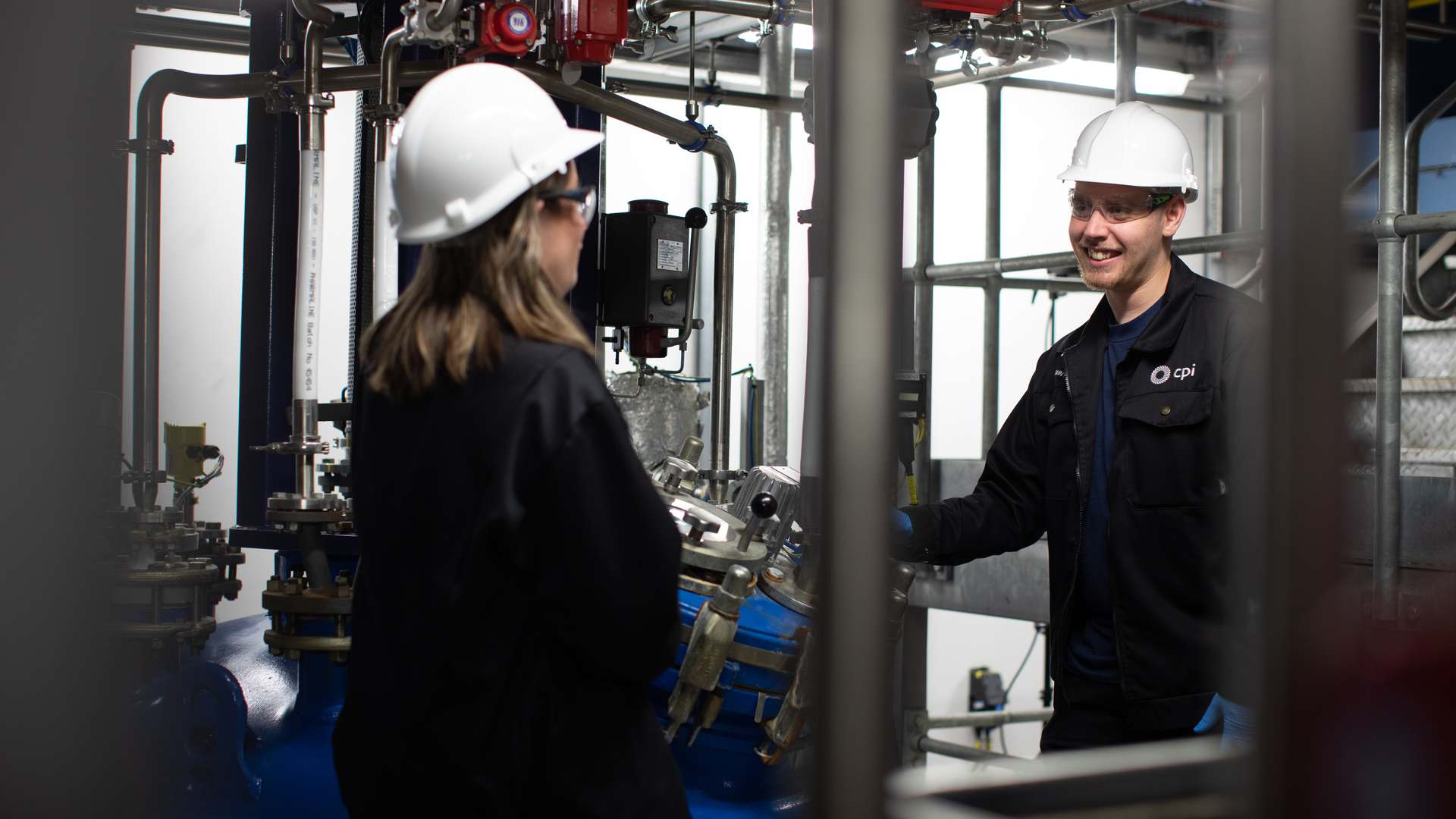 Two of our people in the plant at our Wilton Centre wearing white hard hats talking amongst fermentation machinery. Two of our people in the plant at our Wilton Centre wearing white hard hats talking amongst fermentation machinery.