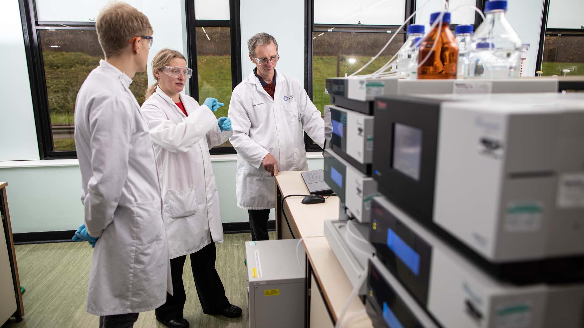 Three of our people in one of our labs at CPI's Wilton Centre wearing white lab coats and goggles standing around a computer at a lab bench and talking. Three of our people in one of our labs at CPI's Wilton Centre wearing white lab coats and goggles standing around a computer at a lab bench and talking.