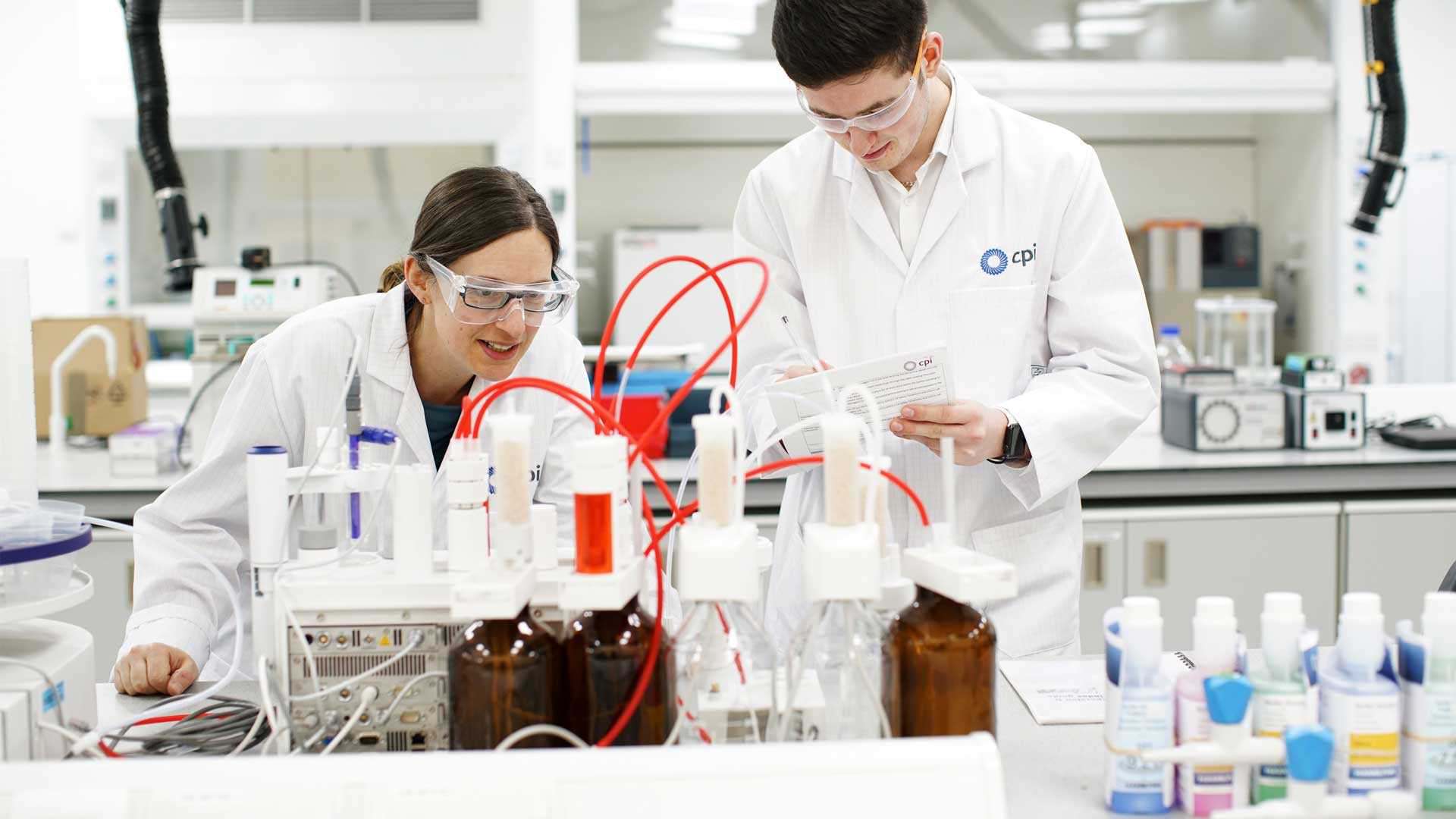 Two CPI scientists wearing white lab coats and googles observe an experiment on a formulations lab bench. Two CPI scientists wearing white lab coats and googles observe an experiment on a formulations lab bench.